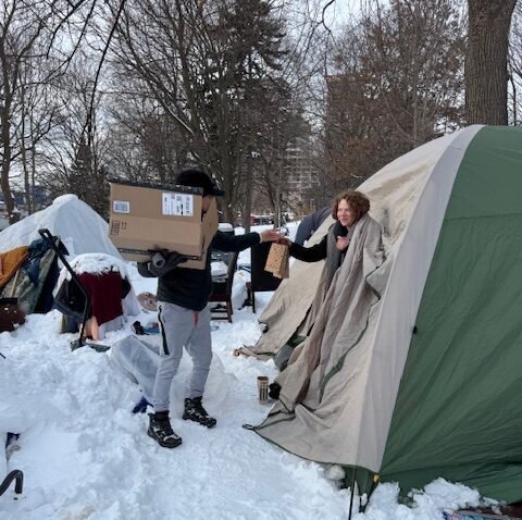 An individual in winter clothing providing direct support and supplies to someone in a snowy outdoor setting.